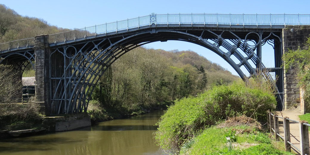 An arched iron bridge spans a calm river, framed by trees and greenery under a clear blue sky.