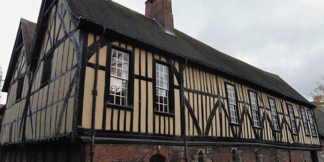 A medieval timber-framed building with yellow plaster infill, large multi-paned windows, and a steep gabled roof with a red brick chimney, likely a guild hall or historic structure.