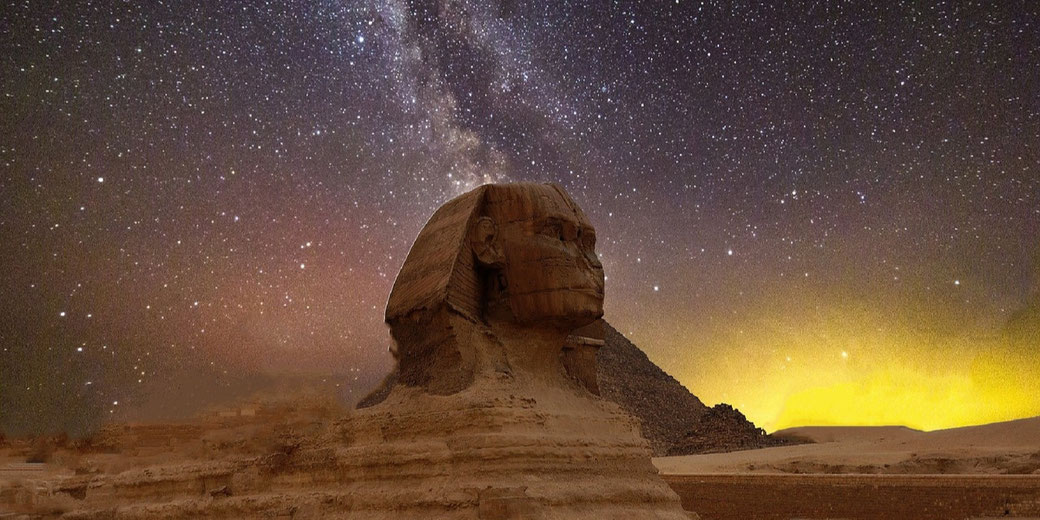 The Great Sphinx of Giza is illuminated under a star-filled night sky with the Milky Way visible above and desert sands surrounding it.