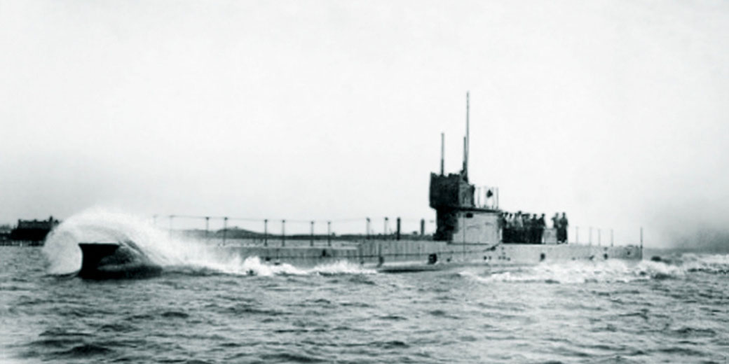 A submarine moves quickly through choppy water, with a group of crew members standing on the deck near the conning tower.
