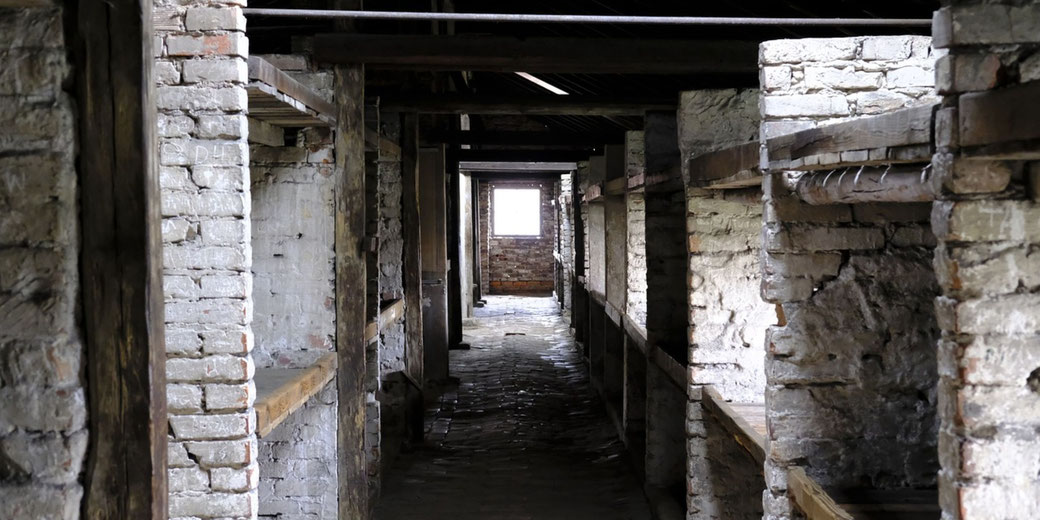 A narrow corridor inside a brick barracks building, with wooden bunks along the walls and light shining through a small window at the end.