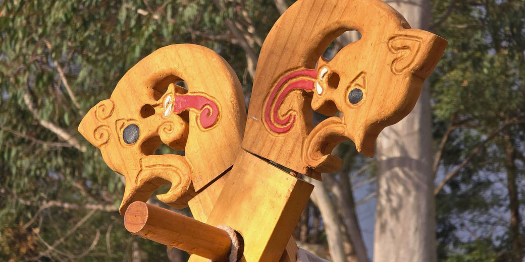 Close-up of two carved wooden animal heads with painted details, mounted on crossed beams against a backdrop of trees in daylight.