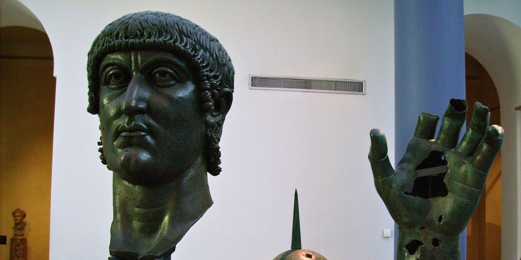 A colossal bronze statue of Emperor Constantine, featuring a detailed head, an outstretched hand with missing fingers, and part of an arm. The fragments are displayed in a museum setting.