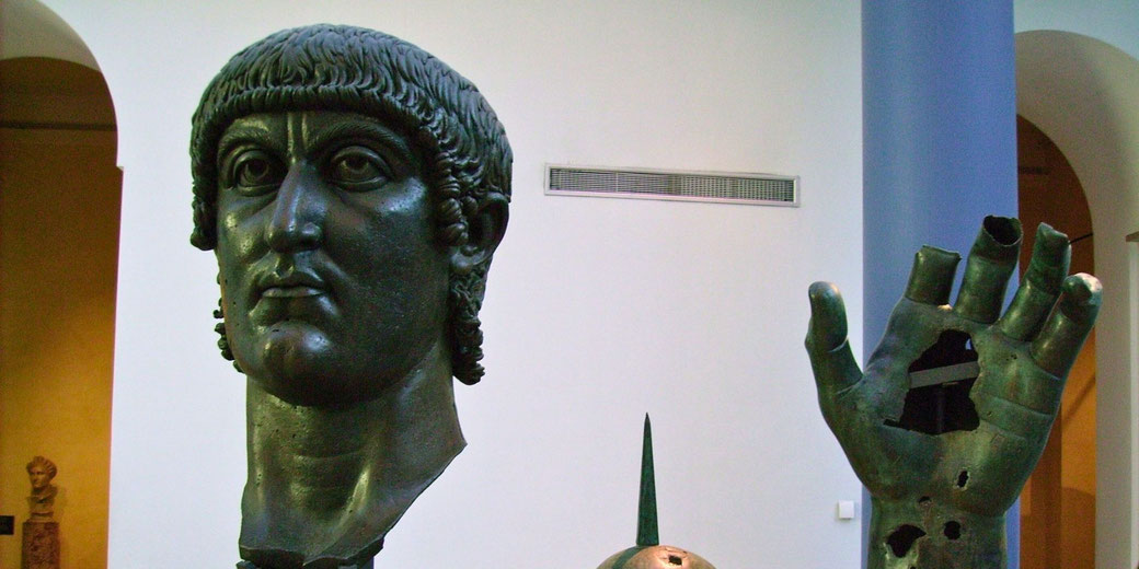 A colossal bronze statue of Emperor Constantine, featuring a detailed head, an outstretched hand with missing fingers, and part of an arm. The fragments are displayed in a museum setting.