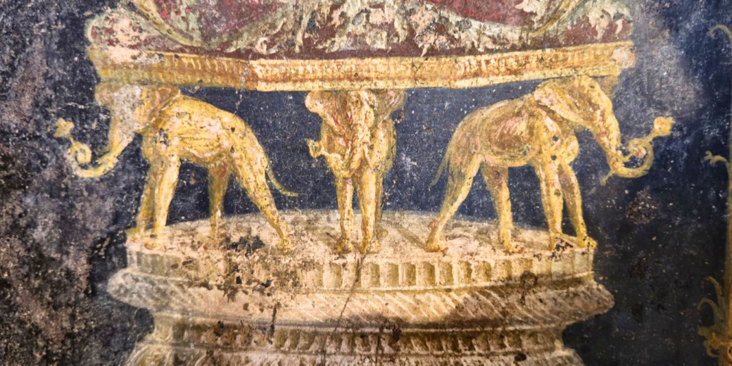 Ancient fresco shows three golden elephants standing on a pedestal, supporting a richly decorated platform above them against a dark background.