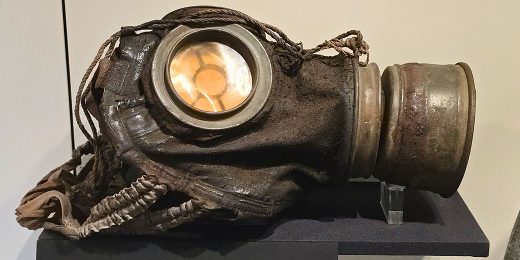 A worn WWI-era gas mask with a large round eye lens, metal filter, and frayed fabric straps displayed on a museum stand.