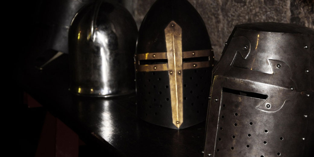 Four medieval knight helmets rest on a dark wooden bench, showcasing different metal designs and visor styles.