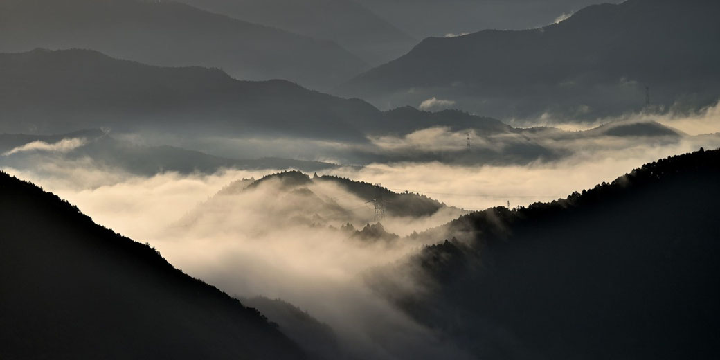 Layered mountain ridges rise through thick fog, with soft light highlighting the mist and silhouettes of power lines faintly visible.
