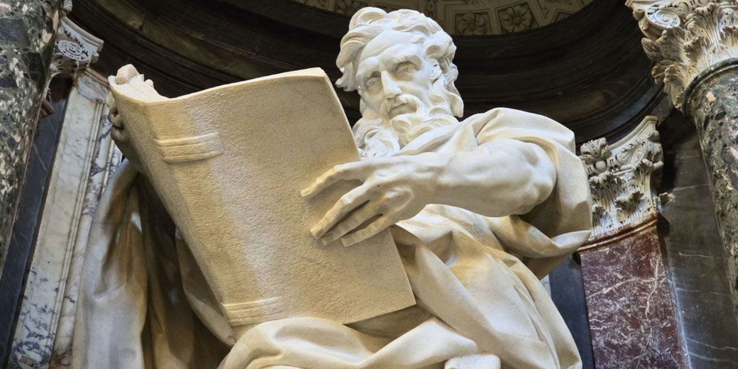 Marble statue of a bearded man deeply focused on a large open book, surrounded by ornate classical columns and drapery.