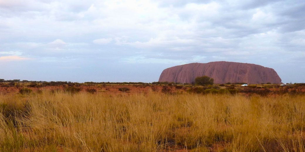 A vast outback landscape with dry grasslands, red earth, and a massive sandstone monolith beneath an overcast sky.