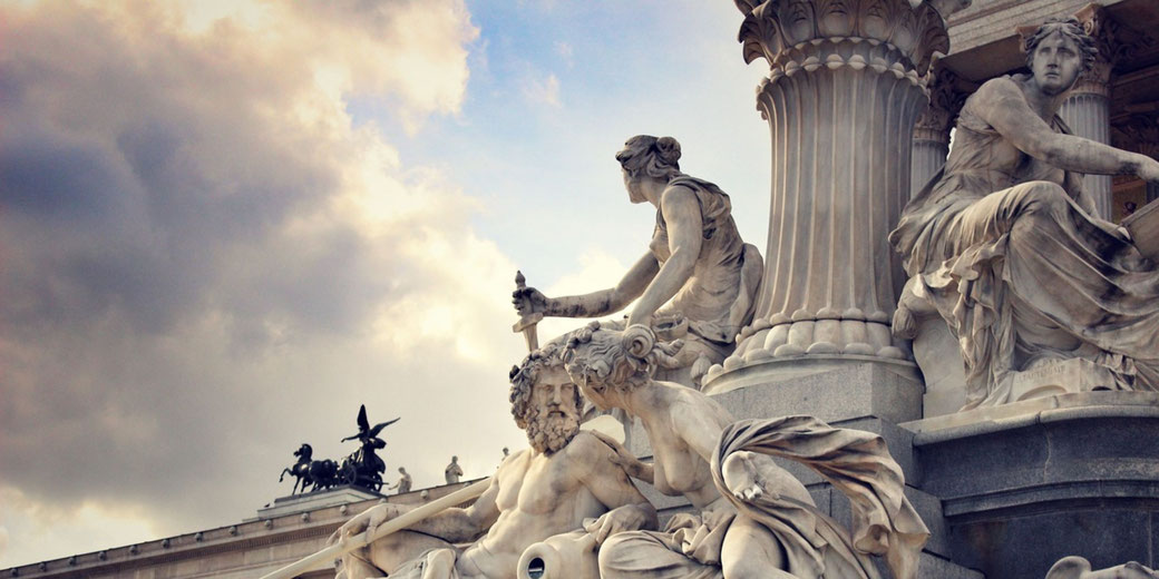 Marble statues of mythological figures with a sword, scroll, and shield surround a tall column; a winged chariot sculpture is visible in the distance.