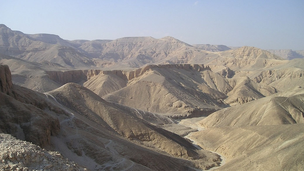 A wide view of barren, rugged desert hills and valleys under a pale blue sky in the Valley of the Kings, Egypt.