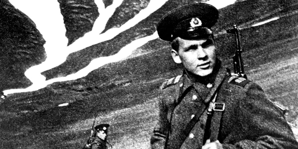 Grainy black and white image of a Soviet soldier in uniform and cap with a rifle slung over his shoulder, standing in a mountainous area with snow-lined slopes.