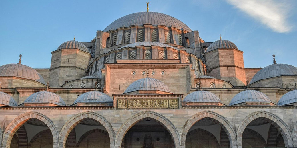 Large historic mosque with central dome and smaller domes, stone arches in front, lit by warm evening light under a clear sky.