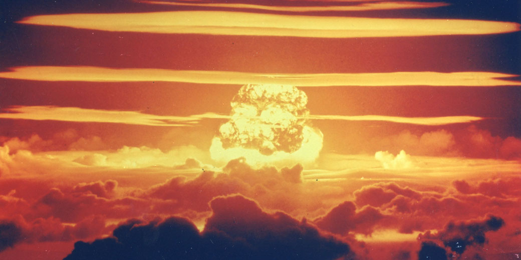A massive nuclear explosion forms a fiery mushroom cloud above the clouds in a dramatic orange sky.