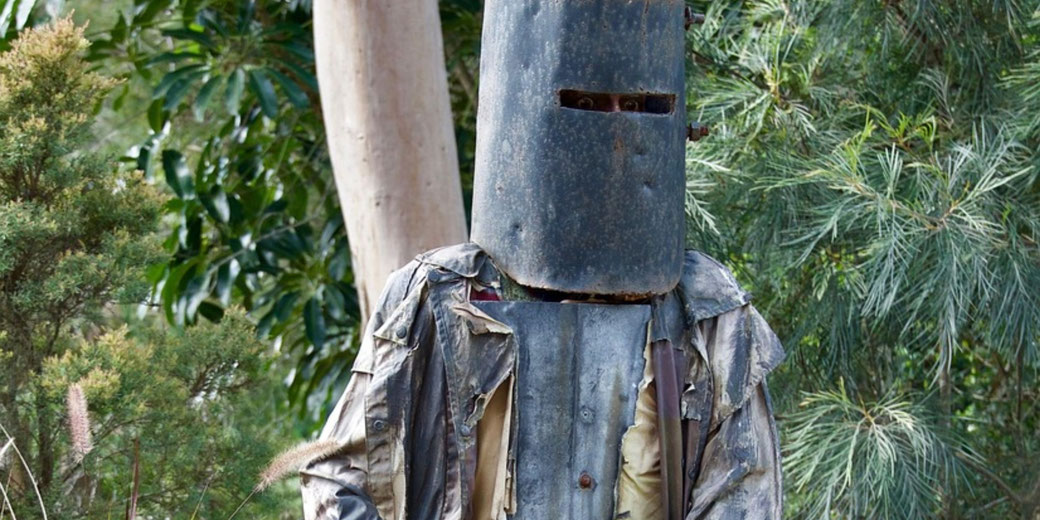 A figure in rusted Ned Kelly armor and ripped coat stands among dense green bushland.