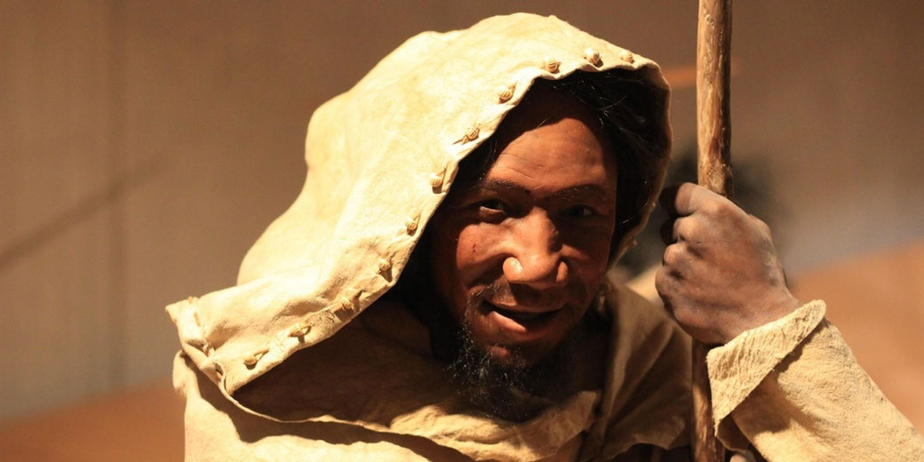 A man in a hooded robe smiles while holding a wooden staff, lit by warm light against a plain background, suggesting a historical or desert setting.