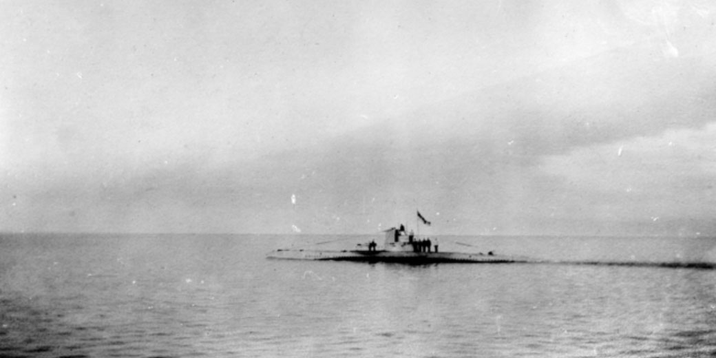 A submarine cruises on the ocean surface with a few crew members visible on deck and a flag flying from the conning tower.