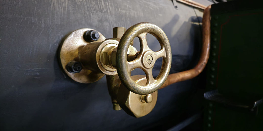 A close-up of a polished brass valve wheel mounted on a dark metallic surface, with copper piping in the background. The industrial design and shiny texture highlight its functional and aesthetic appeal.