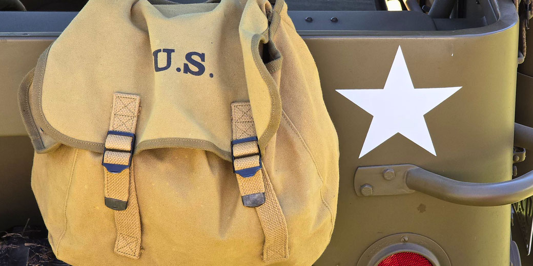 A WWII-era U.S. Army backpack hanging on the back of an olive-drab military jeep, which features a white star insignia. The sturdy canvas gear reflects wartime utility and durability.