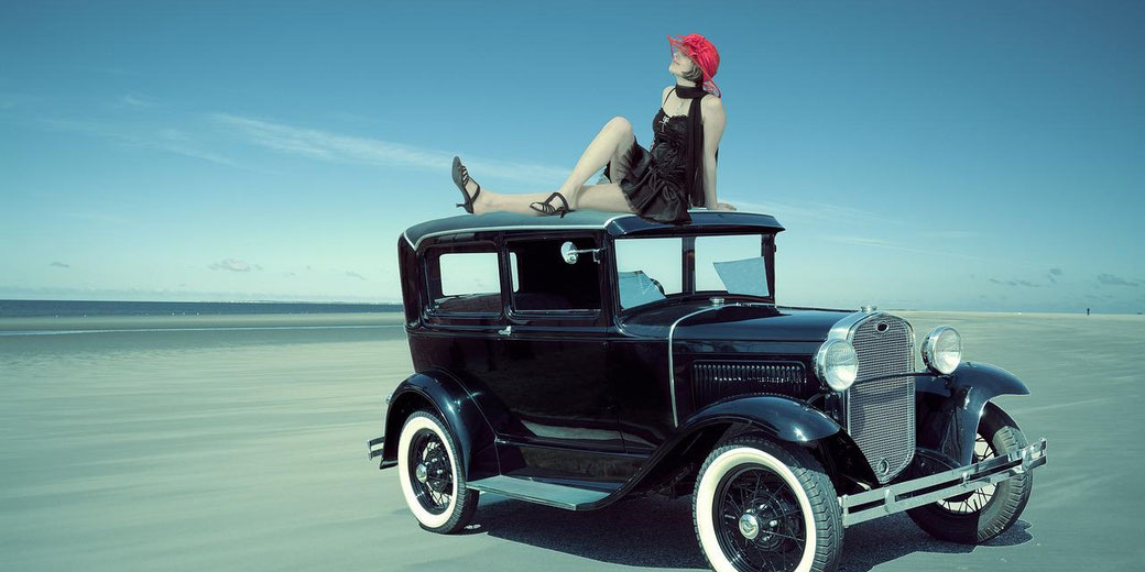 A woman in a black dress and red hat poses on top of a vintage black car parked on a vast, empty beach under a clear sky.