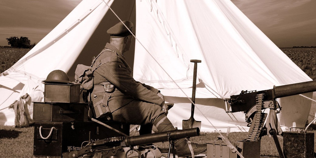 A soldier in vintage uniform sits near a tent with a mounted machine gun and military gear in a sepia-toned scene.