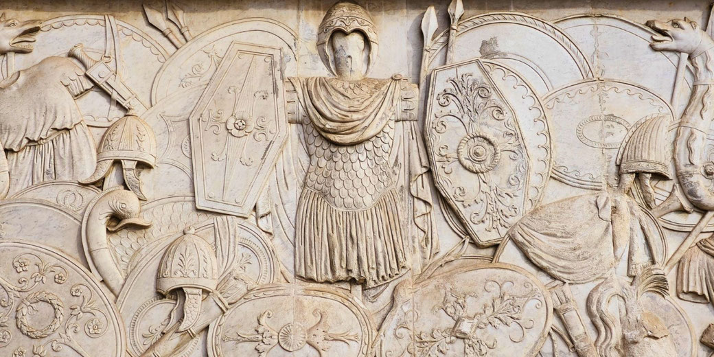 Detailed stone relief of ancient Roman armor, shields, helmets, and weapons arranged around a central cuirass and helmet, symbolizing military valor and triumph.