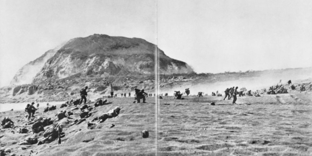 U.S. troops advance on a beach toward Mount Suribachi during the Battle of Iwo Jima, with smoke and sand filling the air.