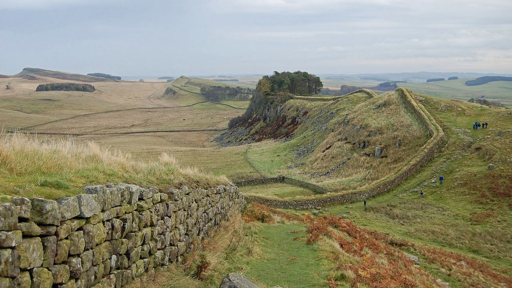 Ancient stone wall winding through grassy hills under a cloudy sky, with small groups of people walking nearby.