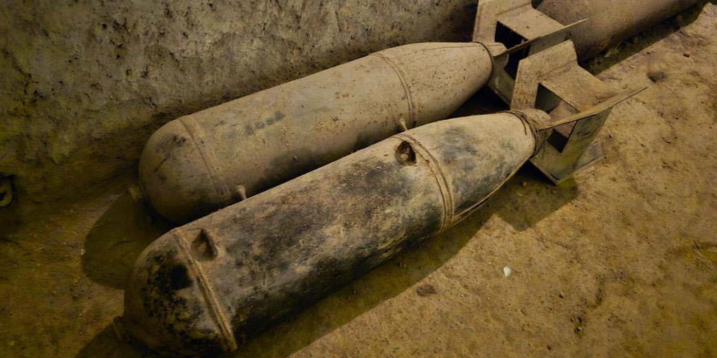 Two large military bombs with tail fins lie on dusty ground inside a bunker-like pit. The metal casings appear aged and dirty, suggesting stored or displayed wartime aerial explosives.