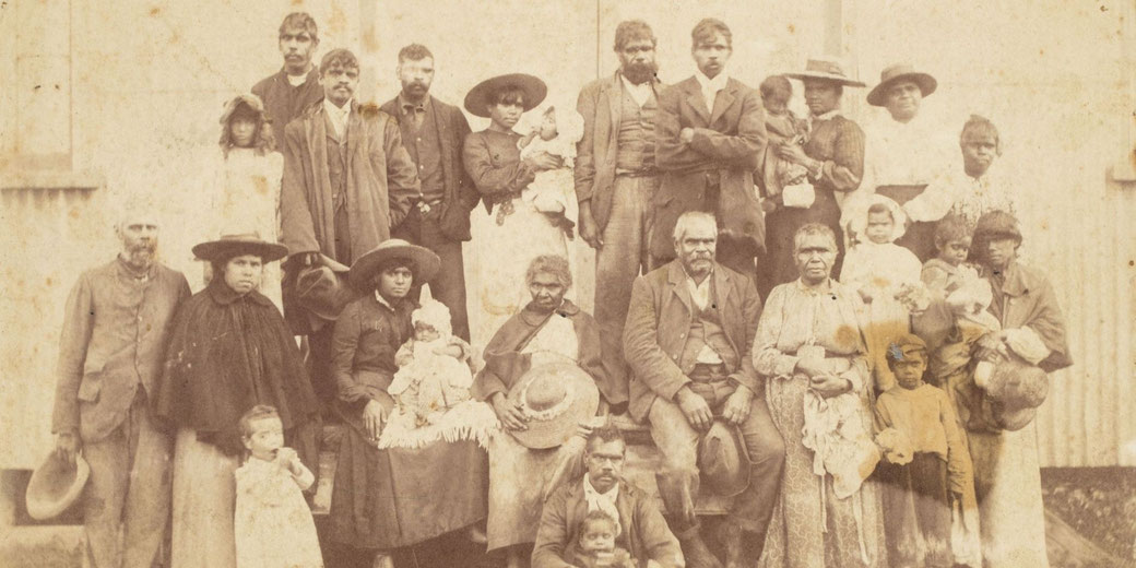 A sepia-toned historical photograph of a large group of Indigenous Australian people, including men, women, and children, posing outside a building.