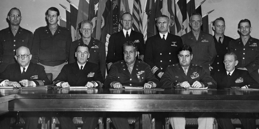 A group of high-ranking military officers poses at a conference table with documents, flags behind them.