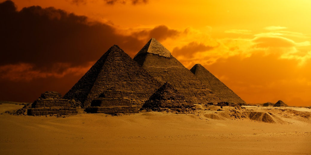 Ancient pyramids of Giza stand in the desert beneath a dramatic orange sky at sunset.