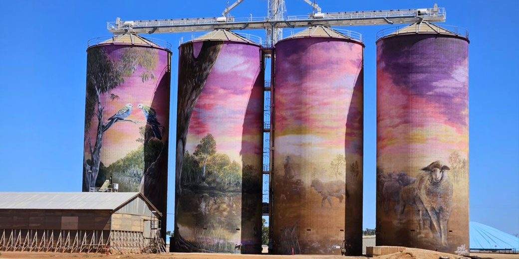 Four tall silos are painted with rural Australian scenes, including parrots, bushland, and sheep under a colorful sky.