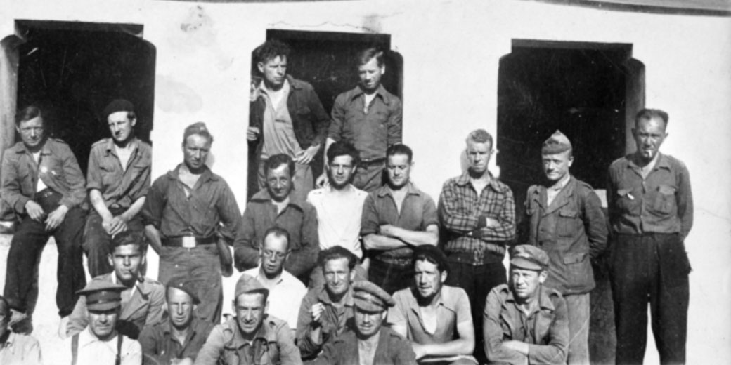A group of men, likely soldiers or workers, pose outside a building in casual and military-style clothing, some standing in window frames, others seated or crouched in front.