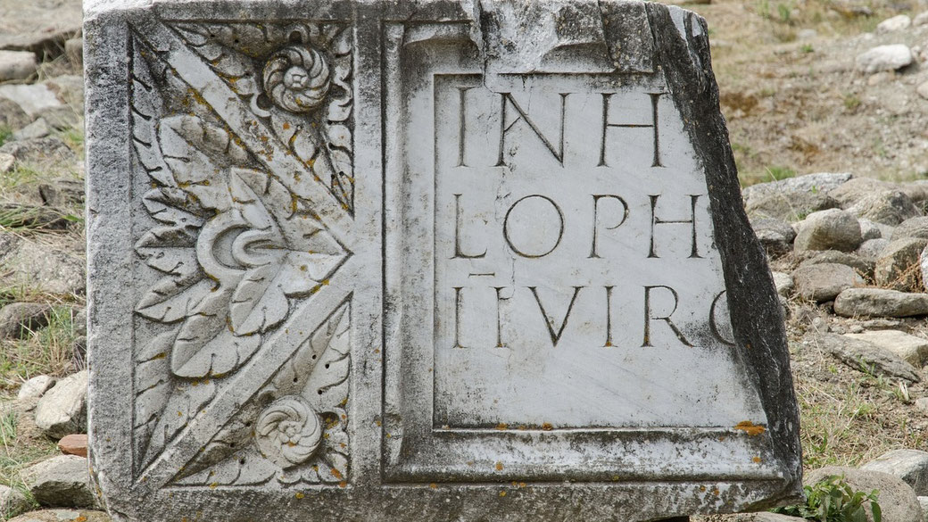 Fragment of a carved Roman marble slab featuring Latin inscriptions and floral relief designs, lying among scattered stones.