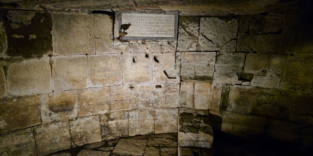 Ancient prison chamber featuring aged limestone walls pockmarked with holes, a mounted Latin plaque, and a stone slab floor showing signs of erosion.