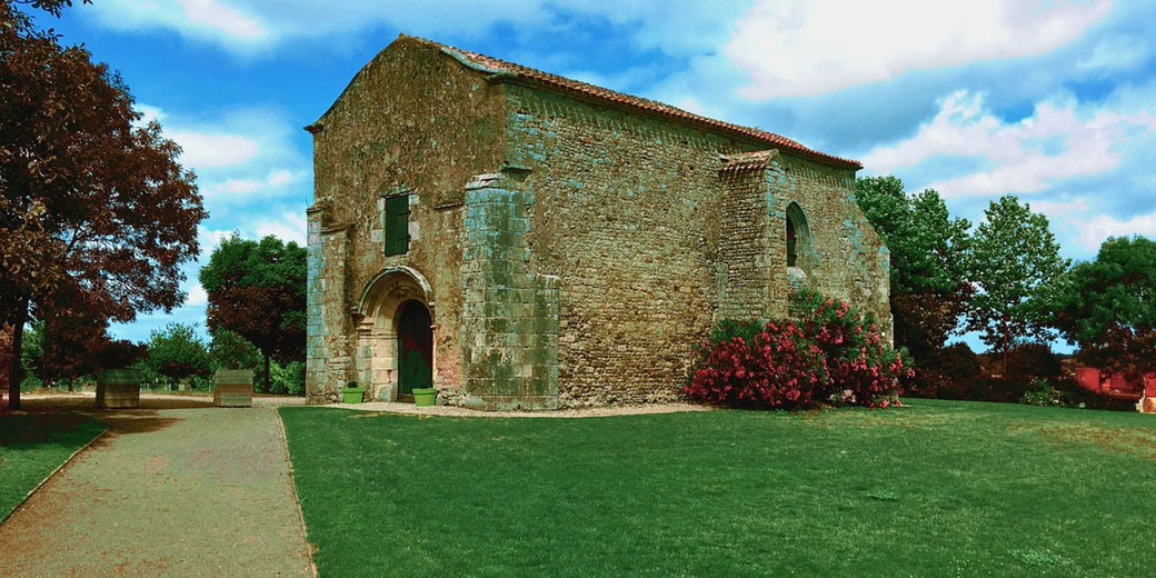 A small, rustic stone chapel stands amid green grass and colorful trees under a bright, partly cloudy sky.
