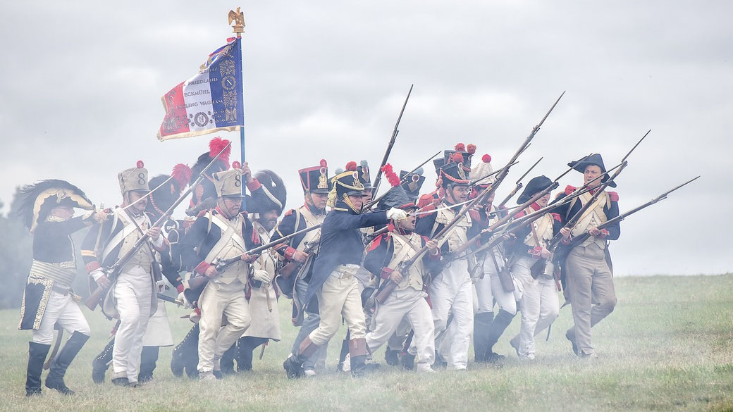 Soldiers in Napoleonic uniforms march forward with raised bayonets and a flag, re-enacting a historical battle scene.