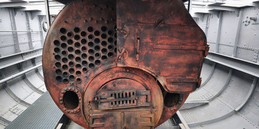 A rusted Escher Wyss boiler inside a ship's interior. The boiler has multiple fire tubes and a furnace door, surrounded by a riveted metal hull, likely part of an old steamship.