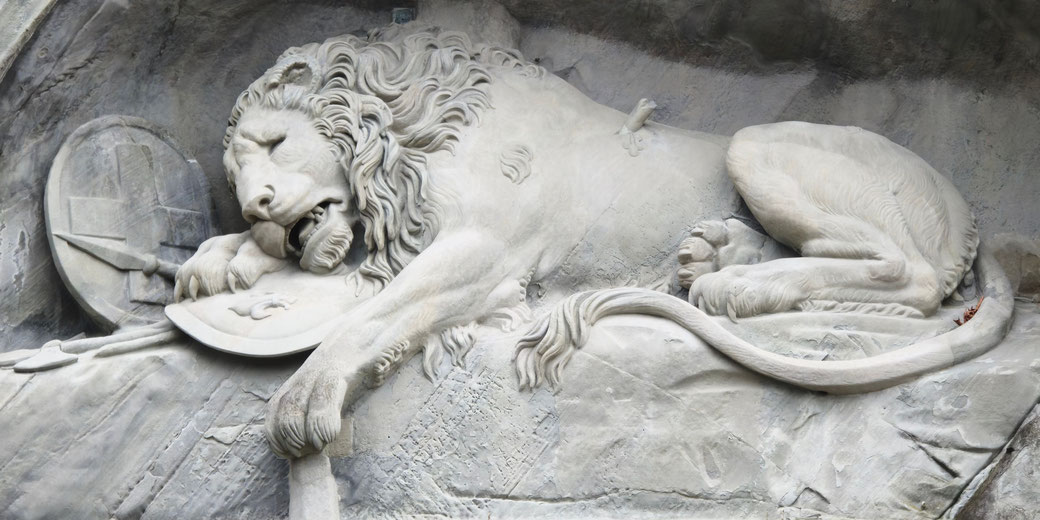 A carved lion lies wounded on a rock, resting its head on a shield with a spear nearby.