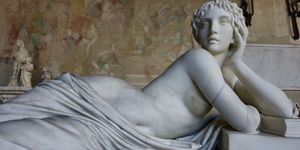 Marble sculpture of a reclining woman resting her head on her hand, gazing upward with a calm expression, surrounded by draped fabric and a faded mural background.