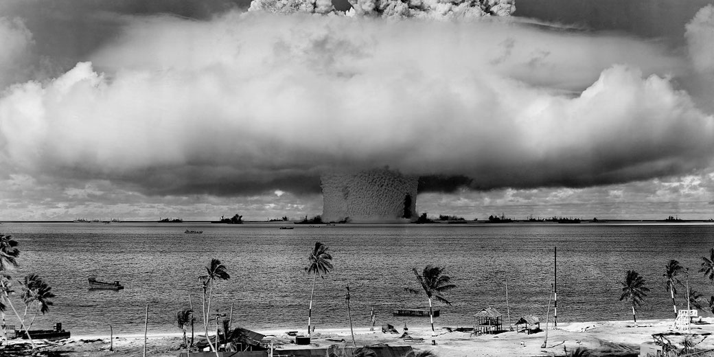 A massive water column rises from a nuclear explosion over the ocean, viewed from a distant tropical shoreline.