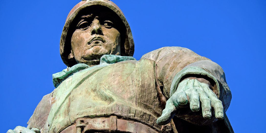 Close-up of a bronze soldier statue in uniform with helmet, looking forward under a clear blue sky.