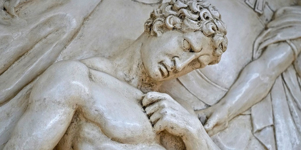 Close-up of a classical relief sculpture showing a muscular male figure with curly hair in a pained or reflective pose.