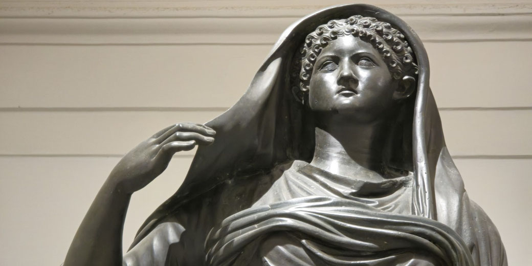 Bronze statue of a veiled woman with curly hair, wearing draped robes and lifting her veil with one hand, exuding calm and dignity.