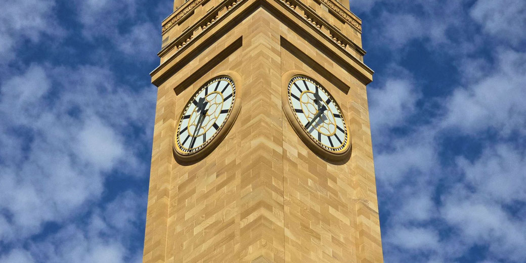 A tall sandstone clock tower with two large round clock faces under a bright sky filled with scattered clouds.