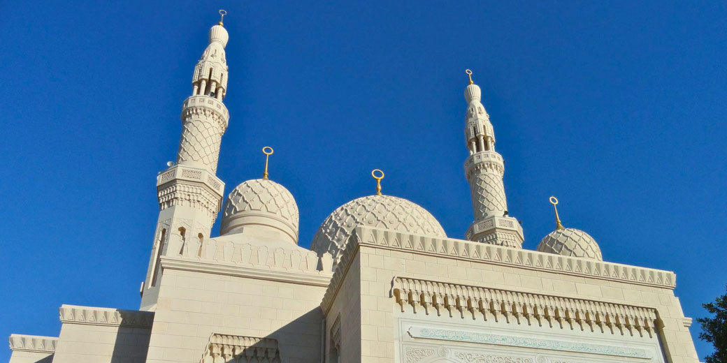 White mosque with patterned domes and tall minarets stands against a vivid blue sky, featuring intricate details and gold crescent finials.