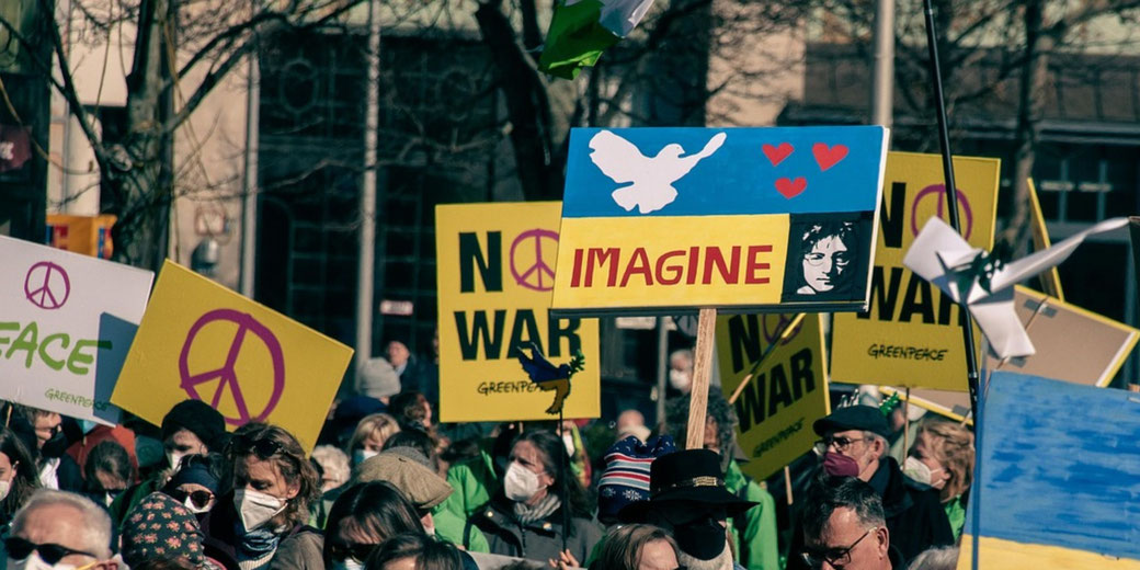 A protest crowd holds peace signs and anti-war posters, including one with John Lennon and the word “IMAGINE”.