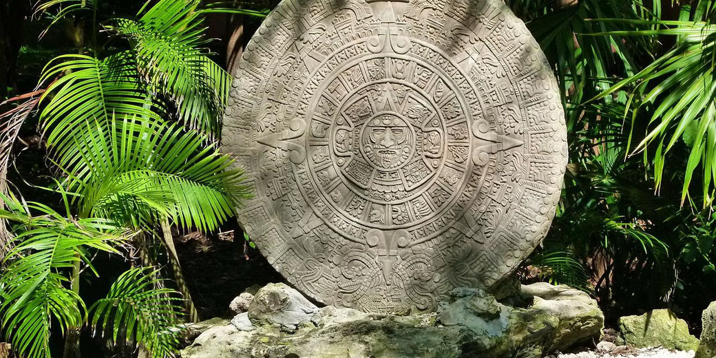 A carved replica of the Aztec Sun Stone, or Calendar Stone, set in a tropical setting with lush green foliage, depicting intricate glyphs and the face of Tonatiuh, the sun god.